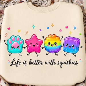 May include: A cream-colored sweatshirt featuring a graphic of four colorful, smiling cartoon shapes holding hands: a teal paw print, a pink star, a rainbow-colored flower, and a blue cube. The text "Life is better with squishies" is printed below.