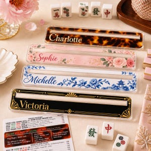 May include: Four rectangular acrylic Mahjong tile racks with horizontal slots. The top rack has a brown tortoiseshell pattern with the name Charlotte. The second features pink roses with the name Sophie. The third has blue floral patterns with the name Michelle. The bottom is black with gold geometric accents and the name Victoria.