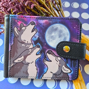 May include: A black and colorful wallet featuring a design of three wolves howling at a full moon. The wallet has a snap closure and a black leather trim. The background is a starry night sky.