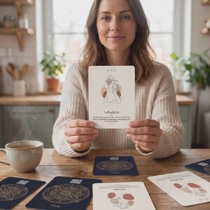 Könnte beinhalten: Eine rechteckige weiße Karte mit einer Strichzeichnung einer Person mit floralen Akzenten und dem deutschen Text 'Selbstliebe', der darunter gedruckt ist. Die Karte wird hochgehalten, weitere Karten und eine Keramiktasse liegen auf dem Tisch.