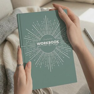 Könnte beinhalten: Ein salbeigrünes Hardcover-Arbeitsbuch mit einer weißen Sonnenstrahl-Grafik in der Mitte des Einbands. Der Text lautet Charlotte WORKBOOK sei wer du bist in weißer und grauer Schrift. Das Buch hat eine glatte, matte Oberfläche und rechteckige Form.