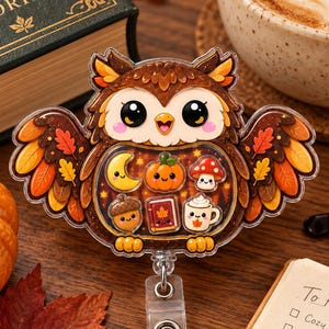 May include: A brown owl-shaped badge reel featuring a clear acrylic front filled with small, colorful icons including a yellow crescent moon, an orange pumpkin, a red mushroom, an acorn, a book, and a white mug with a leaf design.