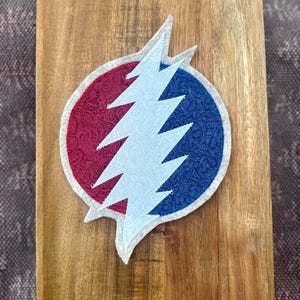 Puede incluir: Un parche de tela circular con un diseño de rayo. El lado izquierdo del círculo tiene un patrón de cachemira rojo, mientras que el lado derecho tiene un patrón floral azul. Un rayo azul claro está centrado sobre el círculo, todo delineado en blanco.