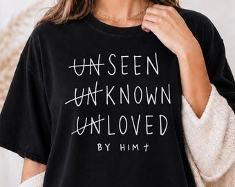 May include: A black short-sleeved t-shirt featuring white text that reads UNSEEN, UNKNOWN, UNLOVED, with each word crossed out by a horizontal line, followed by the text BY HIM and a small cross symbol centered below the words.
