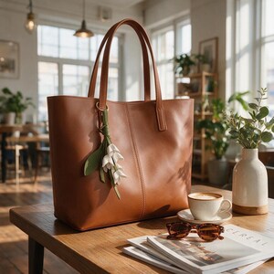 May include: A large, structured brown leather tote bag with two tall shoulder straps. A decorative charm featuring green leaves and white bell-shaped flowers is attached to one of the handle rings. The bag has a smooth finish and vertical stitching.