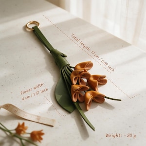 May include: A green leather keychain featuring a gold ring and a cluster of orange flower-shaped charms. The total length is 6.69 inches, the flower width is 1.57 inches, and the weight is approximately 20 grams as indicated in the image.
