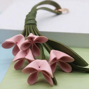 May include: A decorative keychain featuring a cluster of soft pink leather flower blossoms attached to olive green leather stems and a loop. The petals are secured with small pink stitches, and the entire assembly connects to a circular metal ring.