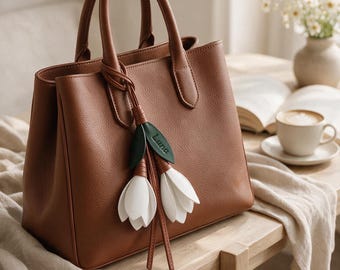 May include: A structured brown handbag with two top handles and a decorative charm featuring two white tulip-shaped pendants with green leaves. One green leaf is embossed with the text Luna. The bag has a textured surface and a clean design.