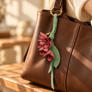 May include: A dark brown leather handbag featuring a decorative charm attached to the handle. The charm consists of several small, textured, dusty rose-colored flower petals and a single light green leaf, all tied with a light green leather strap.