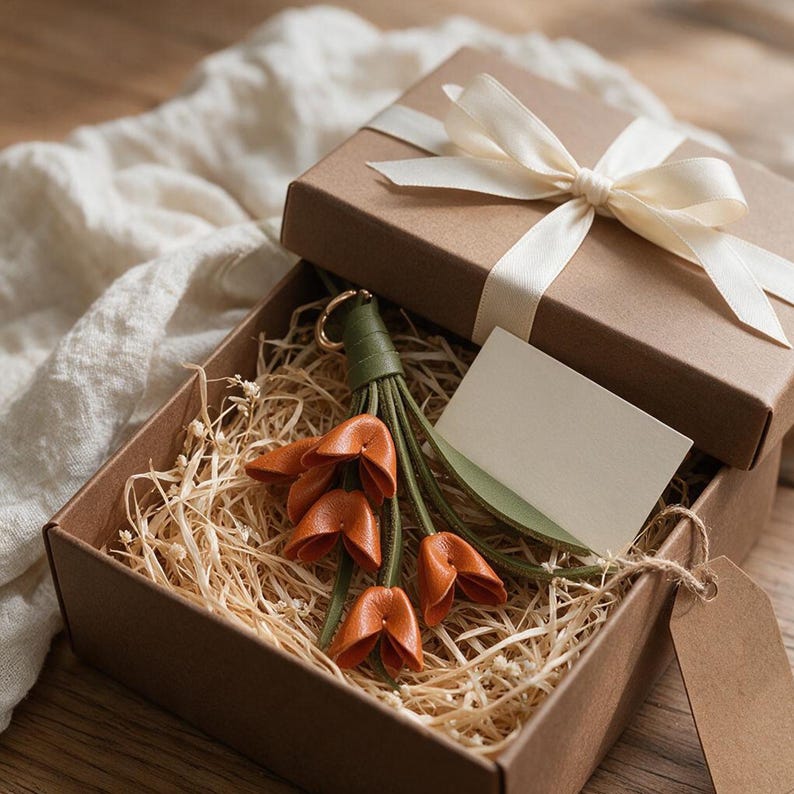 May include: A brown cardboard gift box with a white ribbon contains a leather keychain featuring three orange tulip-shaped charms attached to green stems and a small blank white rectangular card tucked inside the box with straw filler.