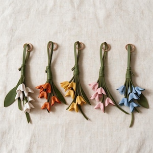 May include: Five decorative leather flower charms, each featuring a green leather strap with a metal ring. The flowers are crafted in white, orange, yellow, pink, and blue, with green leather leaves attached to each stem-like bundle.