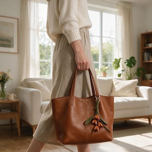 May include: A medium-sized brown leather tote bag with two top handles and a decorative green stem charm featuring three small orange tulip-shaped flowers attached to a gold-toned ring on the front handle base.