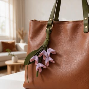 May include: A brown leather tote bag with olive green straps and gold-tone hardware. A decorative charm featuring green leaves and light purple flower petals hangs from a gold ring attached to one of the bag straps near the top zipper closure.
