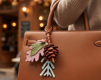 May include: A tan leather handbag featuring a decorative charm attached to the handle. The charm consists of a brown pinecone, a textured green and pink oak leaf, and a light blue sprig, all suspended from a braided leather cord.