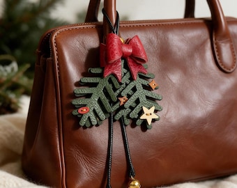 May include: A brown leather handbag featuring a decorative charm shaped like two green pine branches. The charm includes a red leather bow, small wooden star accents, and two dangling black cords finished with small gold-toned metal bells.