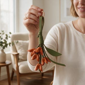 May include: A decorative keychain featuring a cluster of orange leather flower blossoms attached to a green leather strap and a gold-toned metal ring. The design includes several green leather leaves interspersed among the orange petals.