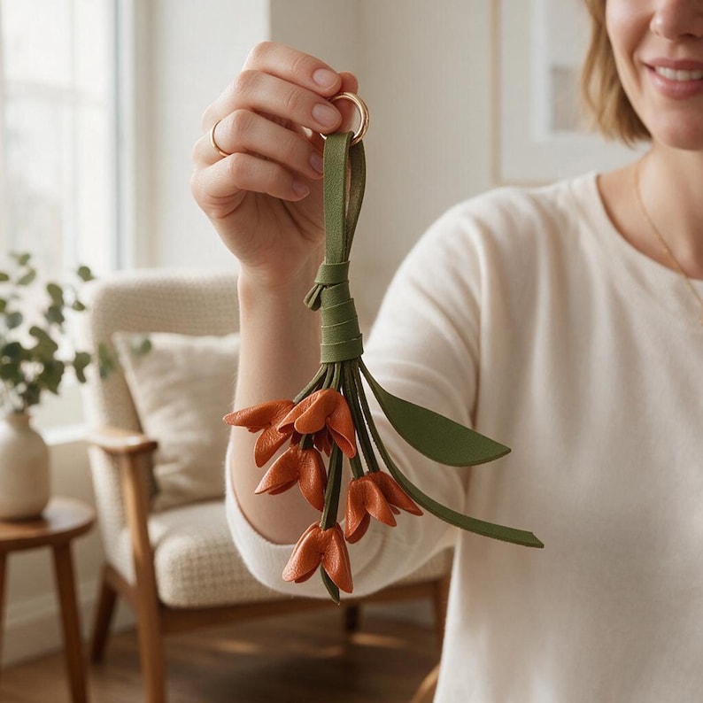 May include: A decorative keychain featuring a cluster of orange leather flower blossoms attached to a green leather strap and a gold-toned metal ring. The design includes several green leather leaves interspersed among the orange petals.