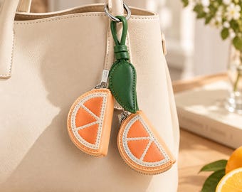 May include: A cream-colored handbag featuring a hanging charm accessory. The charm consists of two orange citrus slice-shaped pouches in bright orange and white, attached to a green leather-like leaf stem with a silver metal ring.