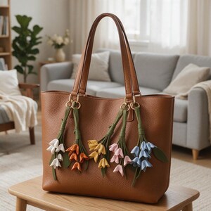 May include: A brown leather tote bag with two shoulder straps, featuring five hanging floral charms in white, orange, yellow, pink, and light blue. Each charm consists of small fabric flowers with green stems and leaves attached to gold rings.