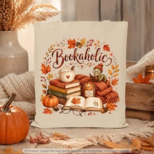 Könnte beinhalten: Eine beigefarbene Stofftasche mit einem aufgedruckten Motiv aus einem Bücherstapel, einer Tasse heißer Schokolade, einem Kürbis, einer Kerze und einem kleinen Wurm mit Brille. Der Schriftzug Bookaholic steht in brauner Schreibschrift darüber.
