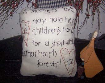 May include: A white pillow with black embroidered text that reads "A mothers love may hold her children's hands for a short while but their hearts forever!" Two red stitched hearts with stars are on the pillow. A wooden angel figurine is to the right.