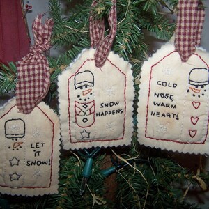 May include: Three handmade Christmas ornaments with snowman designs. Each tag-shaped ornament is cream-colored with red stitching and a red and brown plaid ribbon. The ornaments feature embroidered snowmen and phrases like "Let it Snow!" and "Snow Happens."