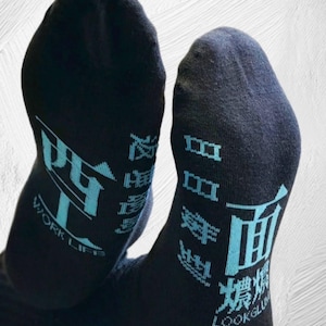 Könnte beinhalten: Ein Paar schwarze Socken mit hellblauer Schrift und Grafiken. Die linke Socke zeigt chinesische Schriftzeichen mit dem Text WORK LIFE, während die rechte Socke chinesische Schriftzeichen mit dem Text LOOK GLUM in einer stilisierten Schriftart aufweist.