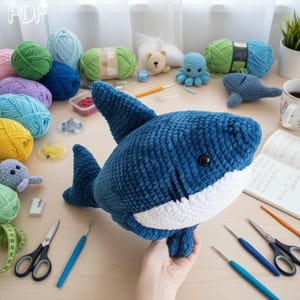 May include: A dark blue and white crocheted shark plush toy with black safety eyes and stitched gill details. The shark is surrounded by various balls of yarn, crochet hooks, scissors, a measuring tape, and other small craft supplies.