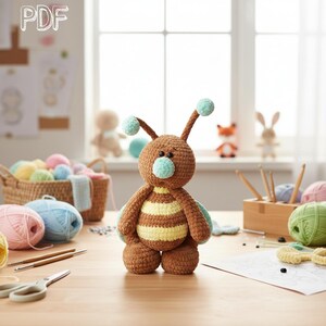 May include: A brown and yellow crocheted bee toy with a light blue nose and two antennae topped with light blue pom-poms. The toy has a round head, a striped body, and two small arms. The text PDF is visible in the top left corner of the image.