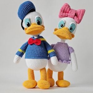 Puede incluir: Dos muñecos de ganchillo hechos a mano que representan patos de dibujos animados. La figura de la izquierda lleva una camisa azul, pajarita roja y gorra azul. La figura de la derecha lleva una camisa morada y un gran lazo rosa en la cabeza.