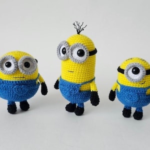 May include: Three crocheted figures featuring yellow bodies, blue overalls, black gloves, and black shoes. The figures have large white and grey goggles with black pupils. One figure is taller with a single tuft of black hair on top.