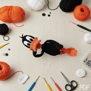 May include: A crocheted black duck doll with a bright orange beak and feet, featuring large white eyes with black pupils. The doll is surrounded by various crafting supplies including orange, black, and white yarn balls, crochet hooks, and scissors.