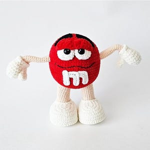 May include: A red, round, crocheted plush toy shaped like a candy character. It features a white letter M on the front, black embroidered eyebrows, black eyes, a stitched smile, beige arms and legs, and white crocheted gloves and shoes.