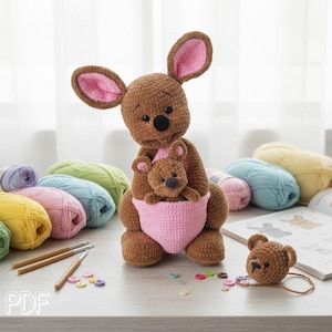Puede incluir: Un juguete de peluche de canguro tejido a ganchillo en color marrón con orejas grandes con detalles interiores rosas, nariz negra y un pequeño canguro bebé dentro de una bolsa tejida de color rosa. Tiene un acabado de hilo suave y texturizado.