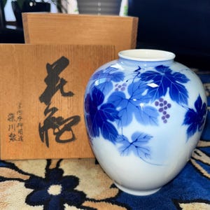 以下が含まれることがあります： 丸みを帯びた胴と細い首を持つ白い陶磁器の花瓶。手描きされた青いブドウの葉と小さな紫色のブドウの房が描かれています。花瓶の横には、黒い日本語の書道と赤い印章が施された木製の収納箱が置かれています。