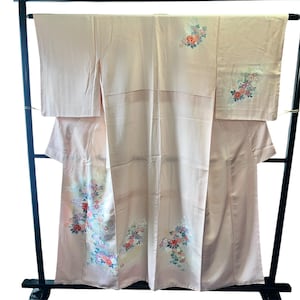 Peut inclure: Un kimono rose pâle orné de délicats bouquets de fleurs dans des tons de rouge, bleu et blanc. Le vêtement présente des manches larges et une base en tissu uni, avec des motifs floraux concentrés sur les panneaux inférieurs et une épaule.
