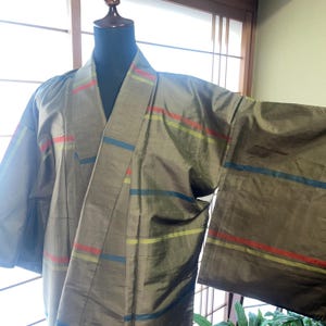 Pode incluir: Uma jaqueta tipo quimono verde-oliva estruturada com um brilho sutil e listras horizontais em vermelho, verde-limão e azul. A peça possui mangas largas, gola larga e design transpassado exibido em um torso de manequim.