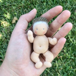 May include: A handmade doll with a beige fabric body, arms, and legs. The doll has a small, round head with white hair and a cap made of a brown material. The doll's face has blue eyes and a small, rosy blush.