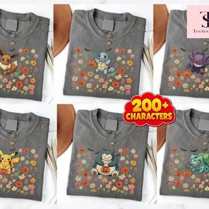 Könnte beinhalten: Eine Kollektion von sechs anthrazitfarbenen T-Shirts, die jeweils eine andere Zeichentrickfigur mit einem kleinen orangefarbenen Kürbis zeigen. Die Motive umfassen Eevee, Squirtle, Gengar, Pikachu, Snorlax und Bulbasaur, umgeben von bunten Herbstblumen.