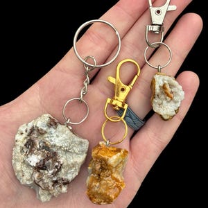 May include: Three keychains featuring raw, natural mineral stones attached to metal hardware. One keychain has a large, textured white and brown stone with a silver ring, another has a golden-brown stone with a gold clasp, and a third has a small white crystal cluster with a silver clasp.