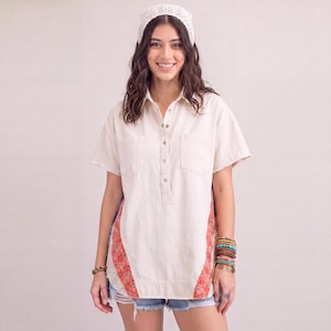 Può includere: Una camicia a maniche corte color crema con bottoni, due tasche sul petto e pannelli laterali con un motivo floreale rosso e blu. La camicia è abbinata a pantaloncini di jeans azzurri e un copricapo all'uncinetto bianco.