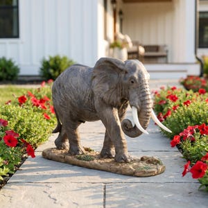 May include: A gray elephant statue with textured skin, large ears, and two white tusks, standing on a brown, uneven base. The elephant is depicted in a walking pose with its trunk curled slightly inward, showing detailed wrinkles and realistic features.