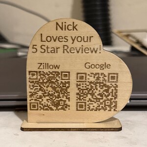 May include: A heart-shaped light wood sign with a flat base featuring the engraved text Nick Loves your 5 Star Review! and two QR codes labeled Zillow and Google. The sign is constructed from a single piece of wood with a dark brown edge finish.