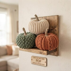 May include: Three decorative pumpkins made of knit fabric in cream, sage green, and burnt orange, resting on a small wooden wall shelf. Below the shelf is a rectangular wooden sign with the text CROCHET PATTERN PDF INSTANT DOWNLOAD.