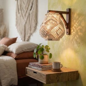 May include: A wall-mounted light fixture featuring a dark brown wooden bracket and a cream-colored macrame shade with intricate woven geometric patterns. The lamp emits a warm glow, casting detailed shadows against the textured light green wall.