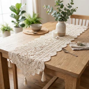May include: A cream-colored crocheted table runner with intricate floral patterns and scalloped edges featuring hanging tassels at the ends. The runner is laid out on a light brown wooden table, accompanied by a white ceramic vase and a small bowl.