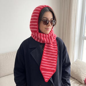 May include: A crochet balaclava hood with an integrated scarf featuring horizontal stripes in alternating shades of bright red and light pink. The headpiece covers the head and neck, with a long scarf tail extending down the front.