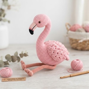 Può includere: Un giocattolo a forma di fenicottero realizzato all'uncinetto in filato rosa chiaro con becco dalla punta nera e occhi marrone scuro. Il fenicottero presenta un design alare testurizzato e zampe lunghe e sottili. Un'etichetta in legno riporta il testo.