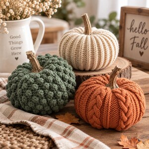 May include: Three decorative pumpkins crafted from yarn in cream, sage green, and burnt orange. The cream pumpkin has a ribbed texture, the green one features a bobble stitch pattern, and the orange one displays a cable knit design with brown stems.