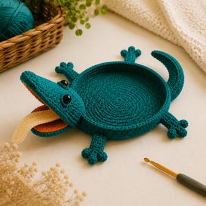May include: A teal crochet lizard-shaped tray with a circular center, four legs, and a curved tail. The lizard has a wide-open mouth featuring a bright orange interior and a long, protruding cream-colored tongue, with two small black bead eyes.